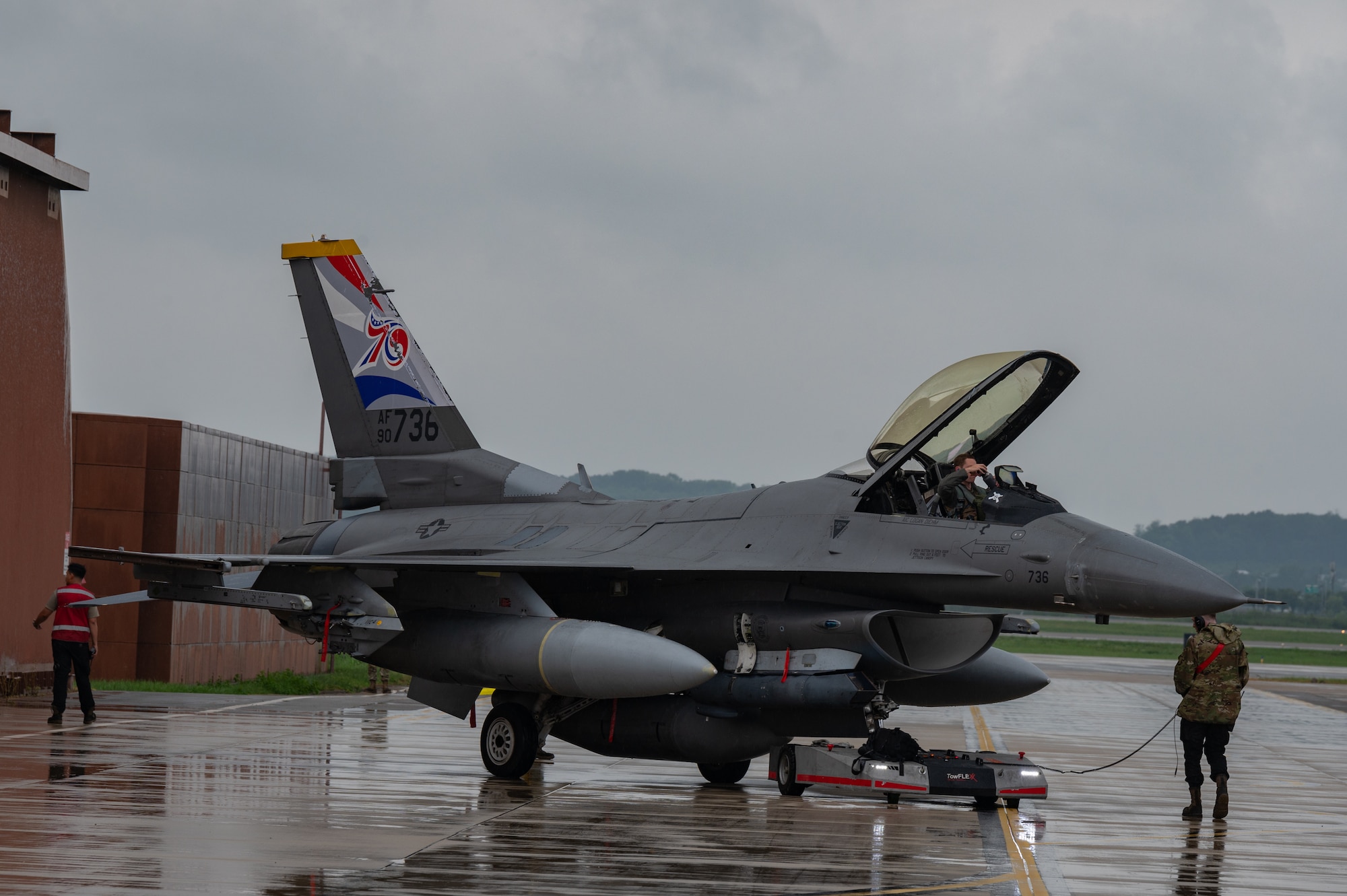 An F-16 fighter jet sits on a wet tarmac facing the camera from the left side of the plane with brown buildings on the left of the photo behind the jet. A person stands with their back to the camera under the front nose of the aircraft.