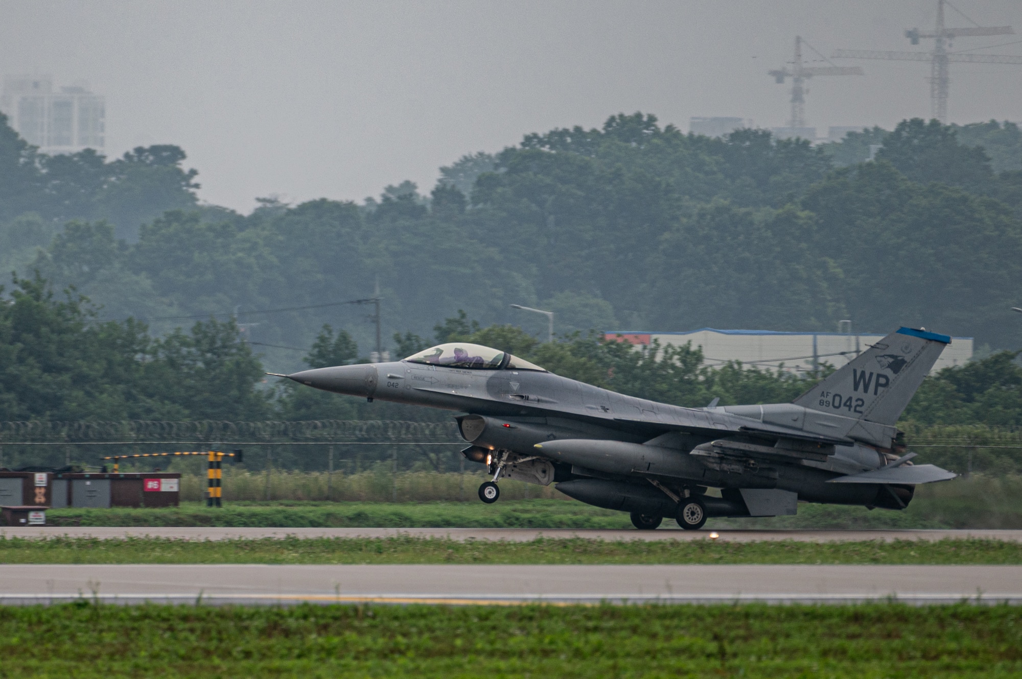 A gray F-16 fighter jet with Kunsan AB WP designation on its tail lands on a runway, viewed from the side profile with the nose pointing left, with green grass and a gray sky. There are trees in the background.