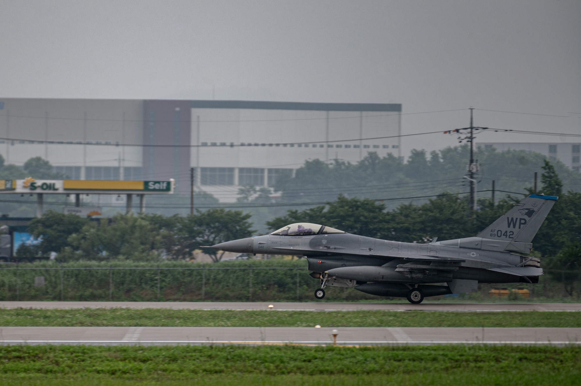 A gray F-16 fighter jet with Kunsan AB WP designation on its tail lands on a runway, viewed from the side profile with the nose pointing left, with green grass and a gray sky. There is a large gray building and trees in the background.
