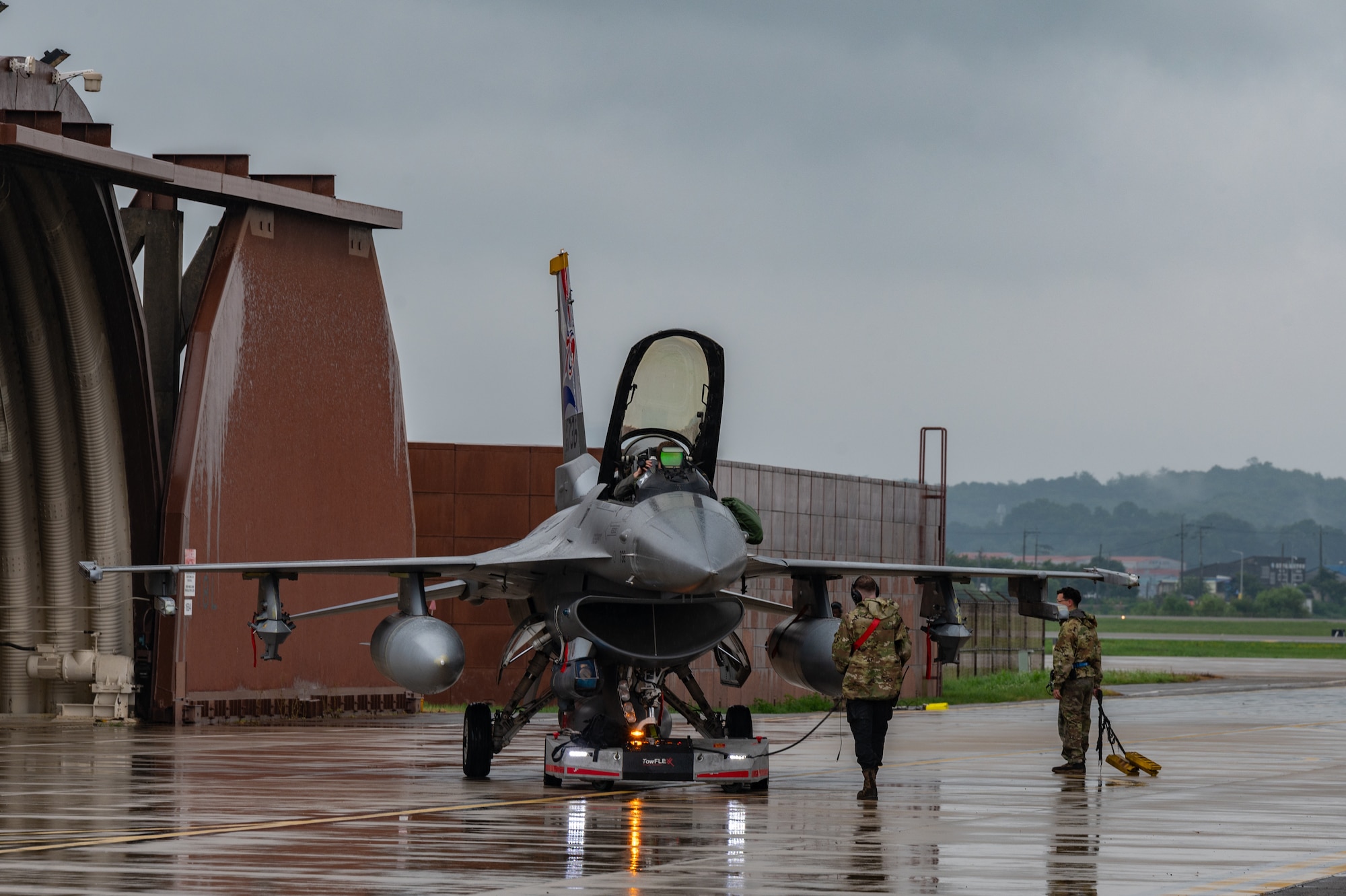 An F-16 fighter jet sits on a wet tarmac facing the camera from the left side of the plane with brown buildings on the left of the photo behind the jet. Two persons stands with their backs to the camera under the right wing of the aircraft.