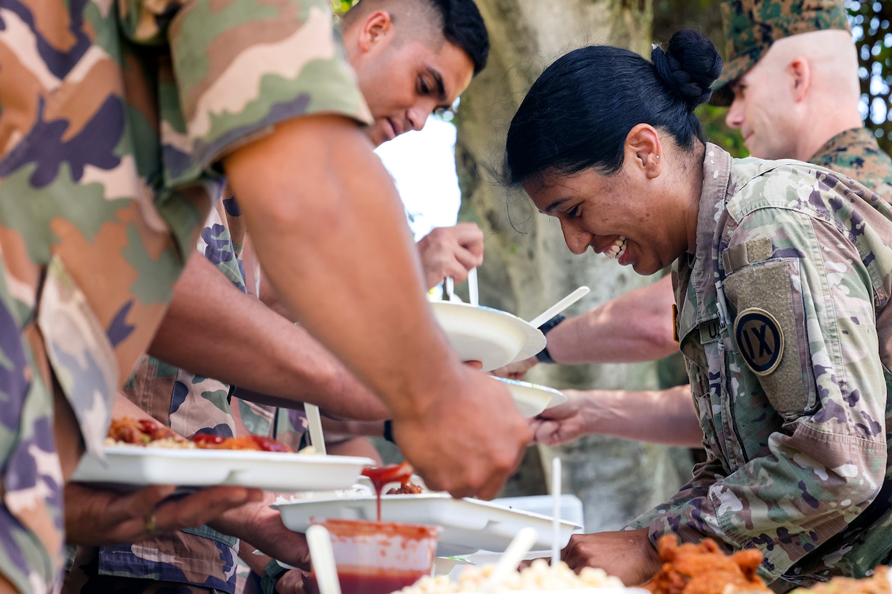 Two smiling soldiers serve other soldiers lunch on paper plates over a table outside.