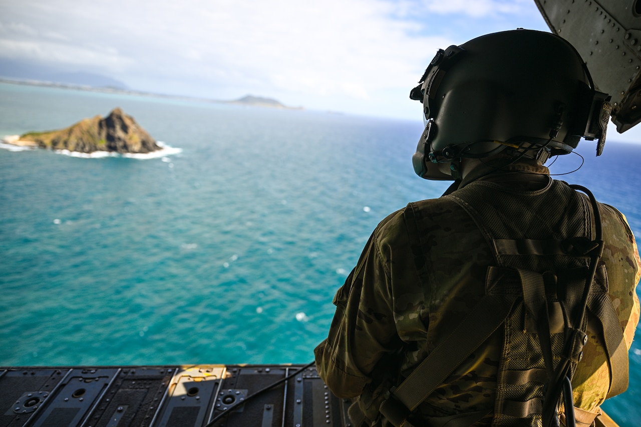 A flight engineer sits on the open deck of a Chinook helicopter looking down on the sea and islands in the distance.