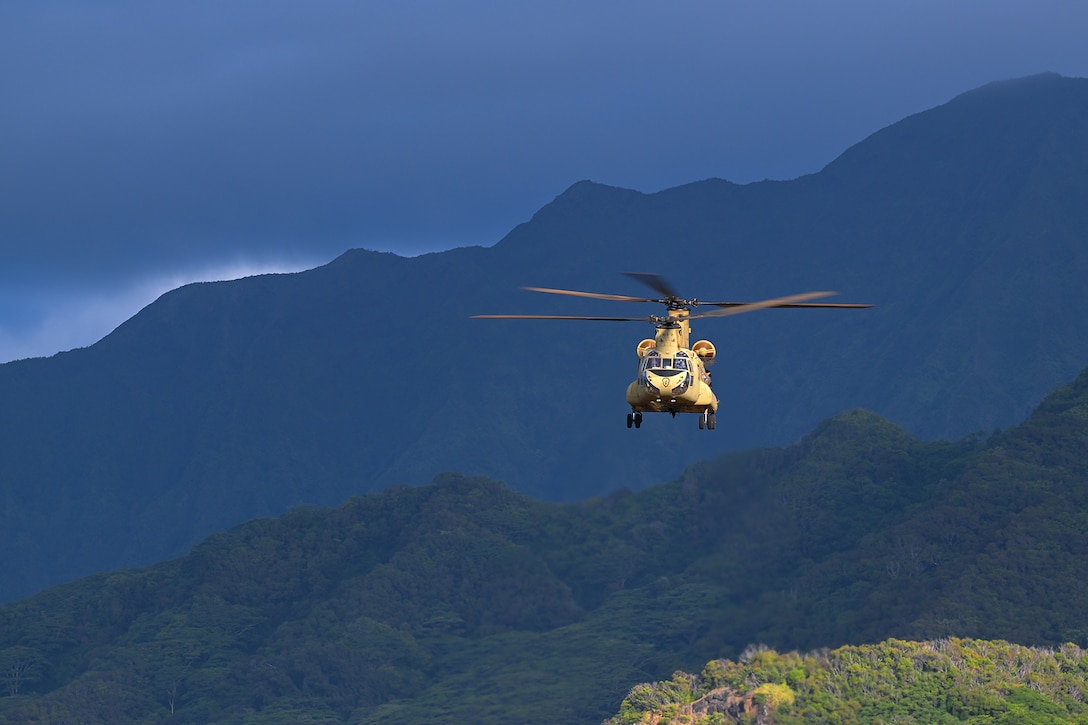 A helicopter flies against a cloudy blue sky during daylight. Mountains and trees can be seen in the background.