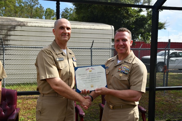 Capt. Marion Gregg, the Commanding Officer of Navy and Marine Corps Force Health Protection Command, presents a Meritorious Service Medal to Cmdr. Ian Sutherland, the off going officer in charge of Navy Entomology Center of Excellence (NECE), at the NECE change of charge ceremony on June 21st, 2024, in Jacksonville, Florida. To commemorate the U.S. Navy’s 248th year of existence, Sutherland had 248 Aedes Aegypti mosquitoes released inside of a seven-foot cube to join him during his speech. To NECE’s knowledge, this is the first-time mosquitoes have been utilized in a change of charge ceremony. (Courtesy photo by Mass Communicator Specialist 2nd Class Matthew Riggs)