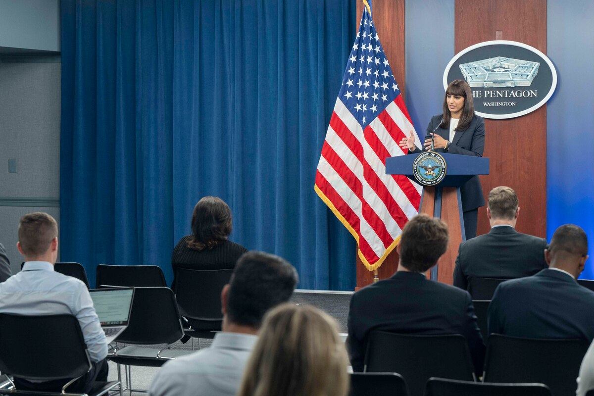 A woman stands behind a lectern with people seated in front of her. A sign on the wall indicates that this is taking place at the Pentagon.