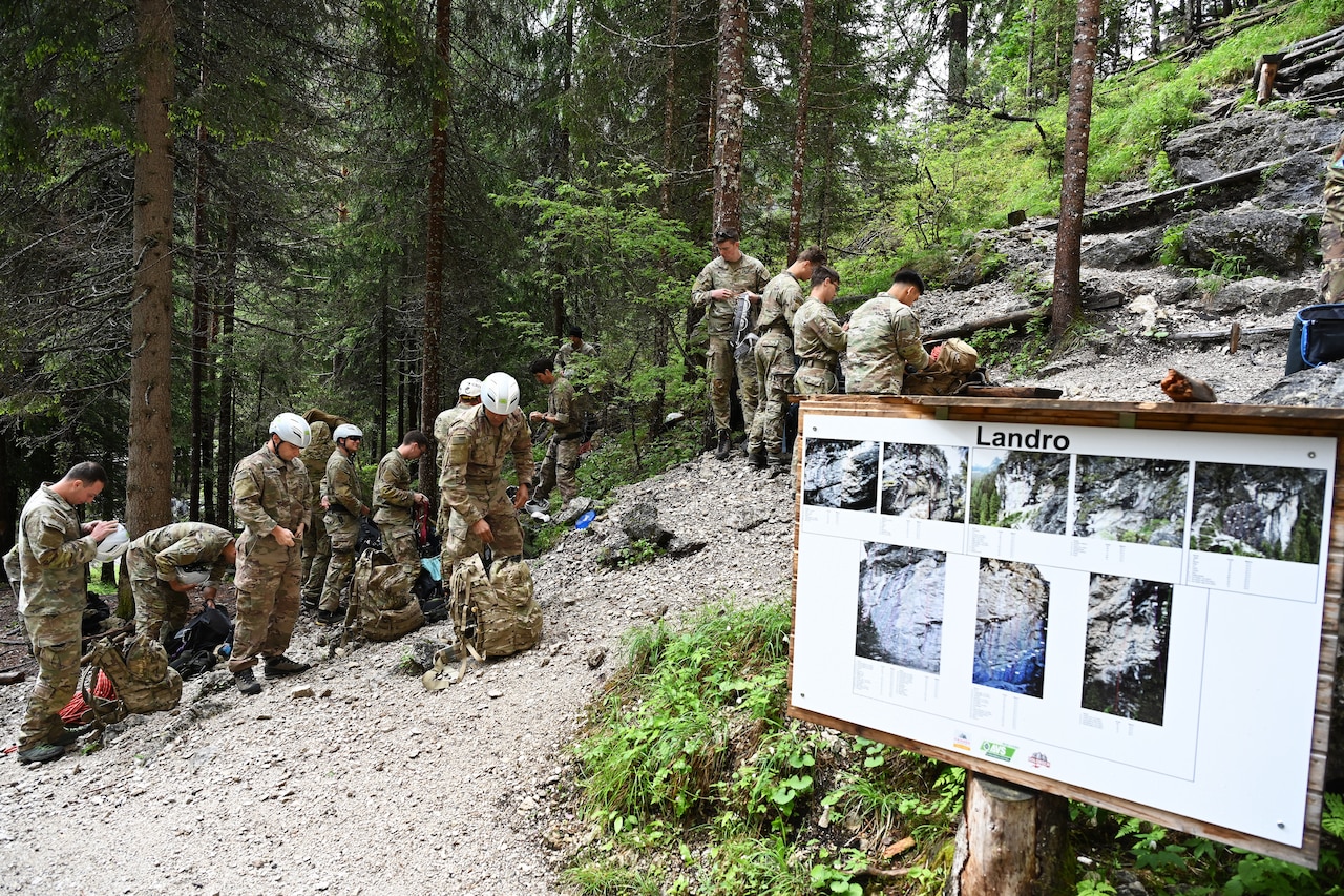 A group of paratroopers puts on climbing gear while on a mountain trail sorrowed by trees and greenery.