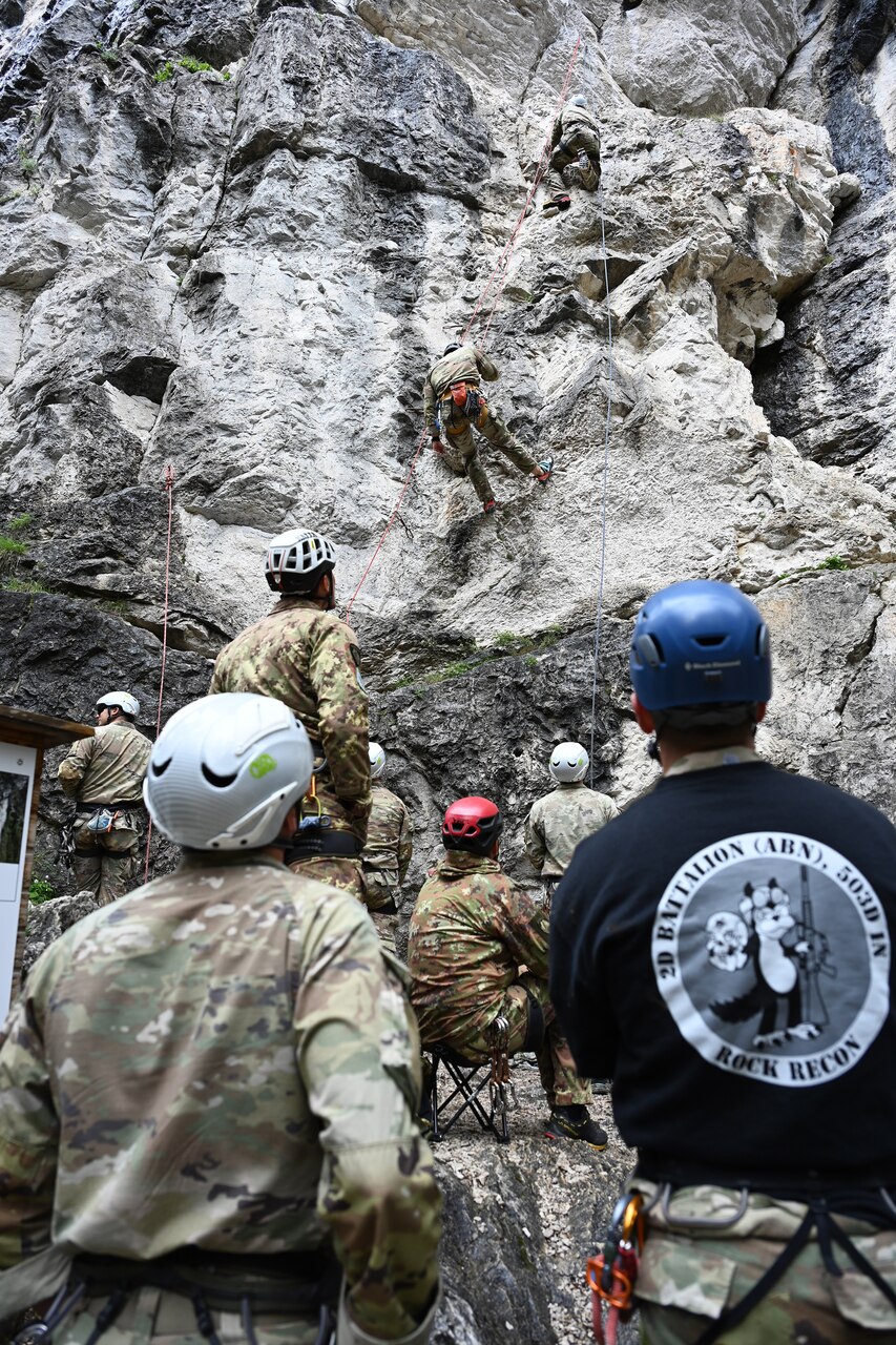 Two soldiers with climbing equipment grapple up a mountain face while other participants look on from the bottom.