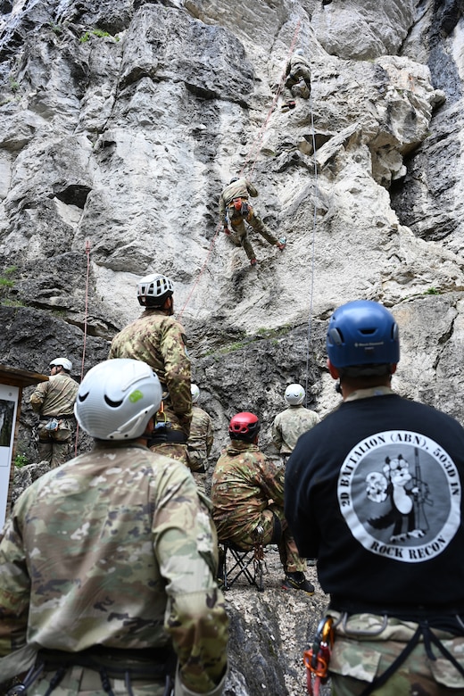 Two soldiers with climbing equipment grapple up a mountain face while other participants look on from the bottom.