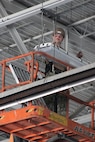 A Seabee conducts maintenance on a lightning fixture in a hangar bay.