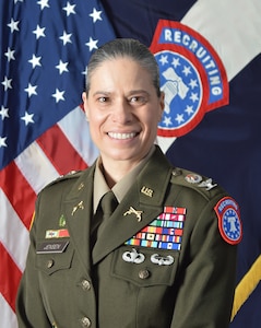 Female Army Soldier in uniform with medals smiles while posed in front of both the U.S. and the Army Recruiting Brigade flags
