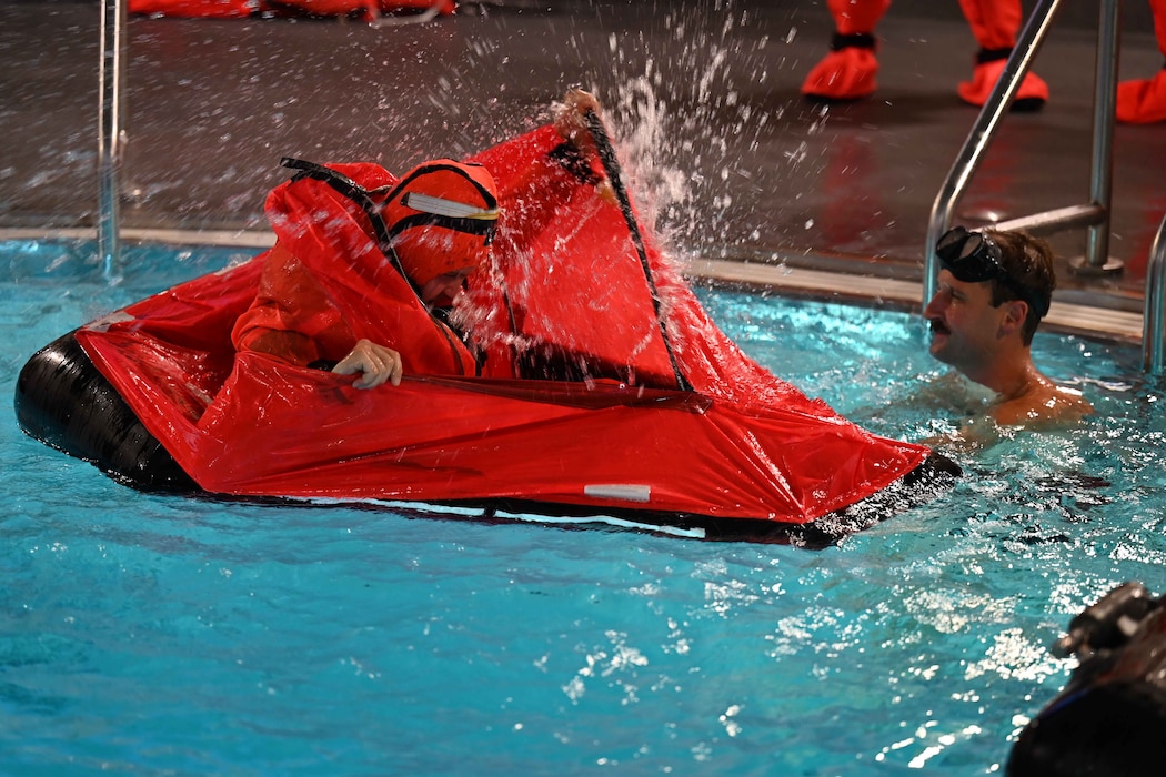 A submarine school student surfaces in a red floating tent raft in a swimming pool as another student looks on and smiles.