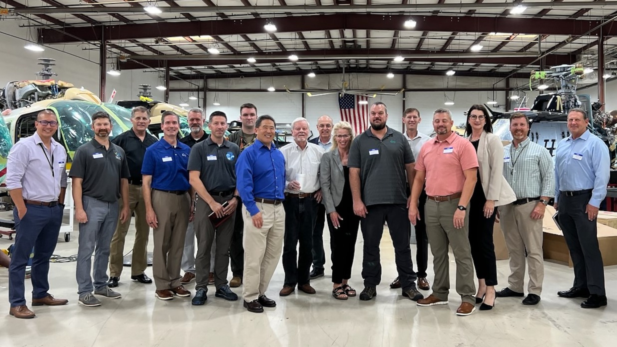 Seventeen people stand together to pose and smile for a group photo. They are located inside of an aircraft hangar. Their background shows helicopters in various stages of maintenance with a large American flag on the wall at the very back, which can be seen behind the heads of the people in the group photo.
