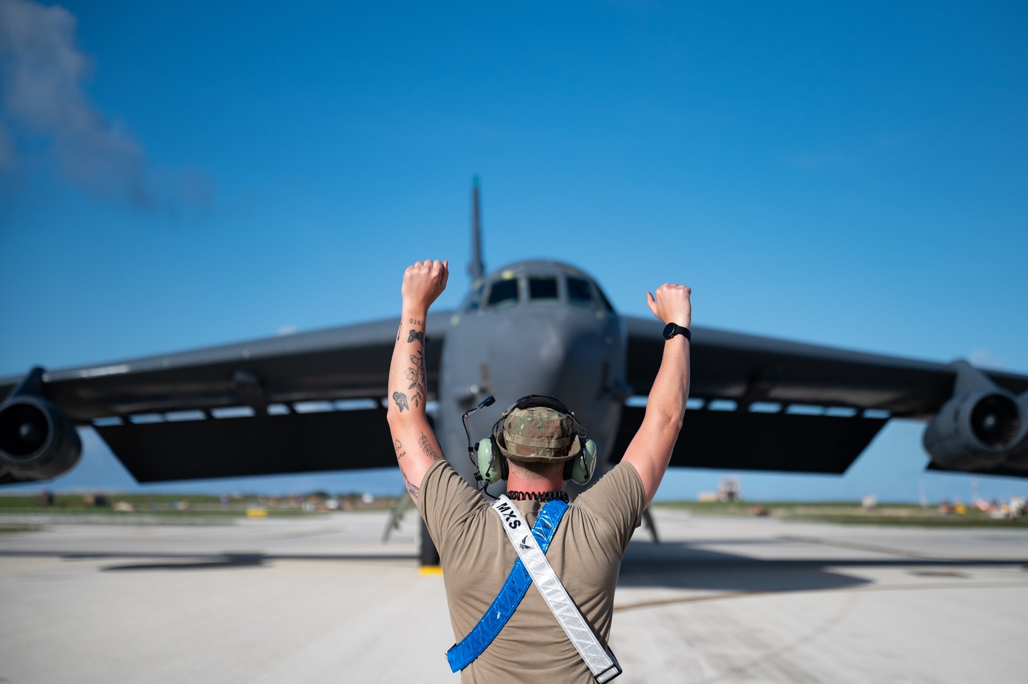 Overall location is outside on a flightline with blue skies. The perspective is from someone who is standing on the ground behind a man, so the image shows the back of a man who is also standing on the ground and is holding up both of his arms in the air with his hands making fists. There is a large, gray military aircraft, specifically a B-52 Stratofortress, also on the ground that is facing the man and cameraperson, the man with arms in the air is has his arms positioned like this to signal to the aircraft pilots to bring the aircraft in front of him and park there.