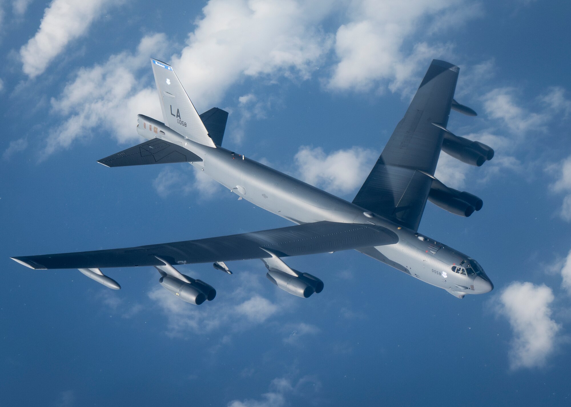 The overall location is in the air, with the photographer's perspective being from inside an aircraft. The photo is of a B-52 Stratofortress assigned to the 2nd Bomb Wing, Barksdale Air Force Base, Louisiana, is flying in the air. The sky is blue with white clouds around it. The aircraft is a large, gray military style. The composition of the picture has the aircraft nose pointing towards the right, so the entire wingspan is on display and the initials "LA" for Louisiana are seen on the tail.