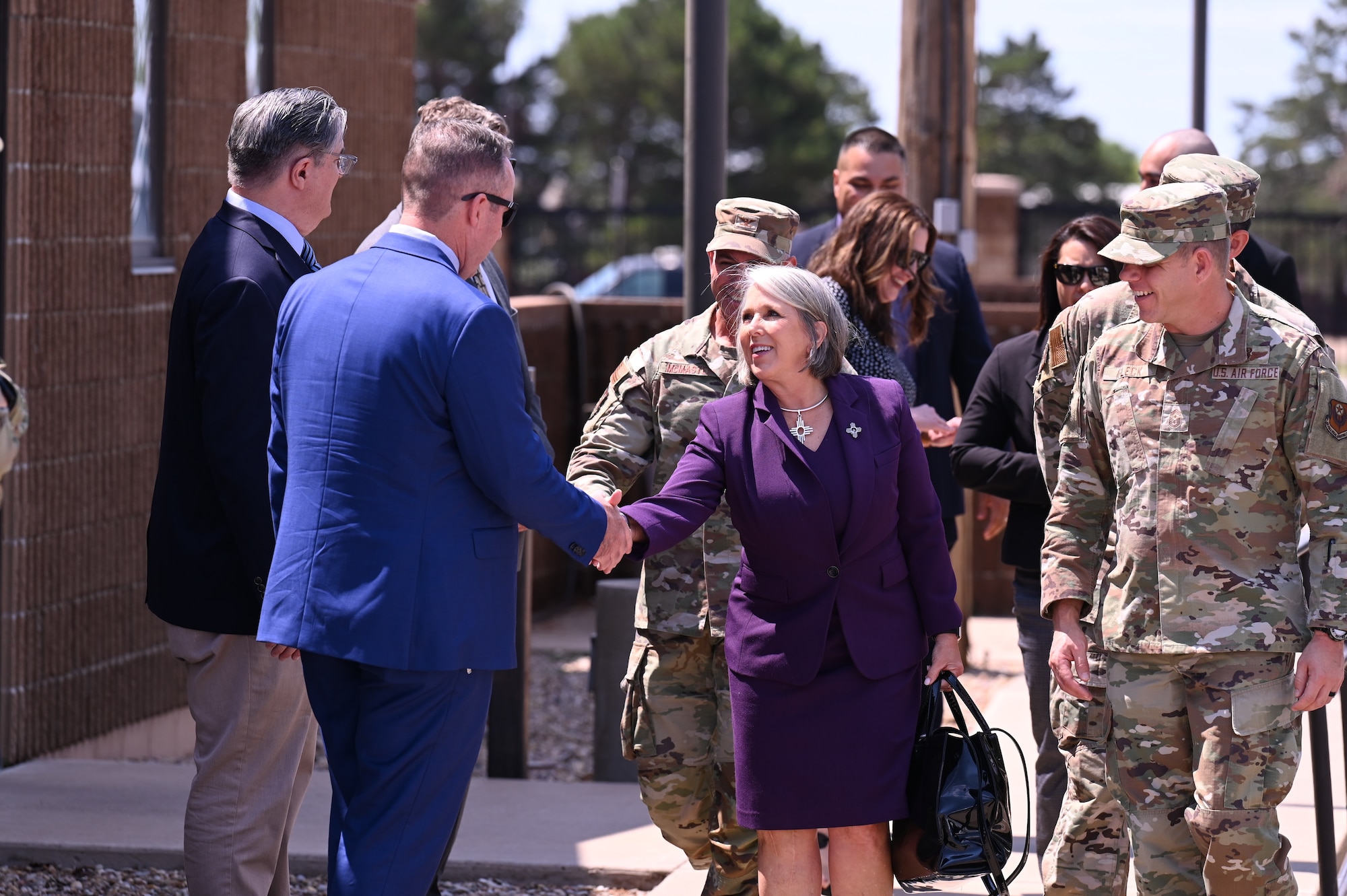 A woman in business attire shakes hands with a man in business attire while a group of men in military uniform look on.