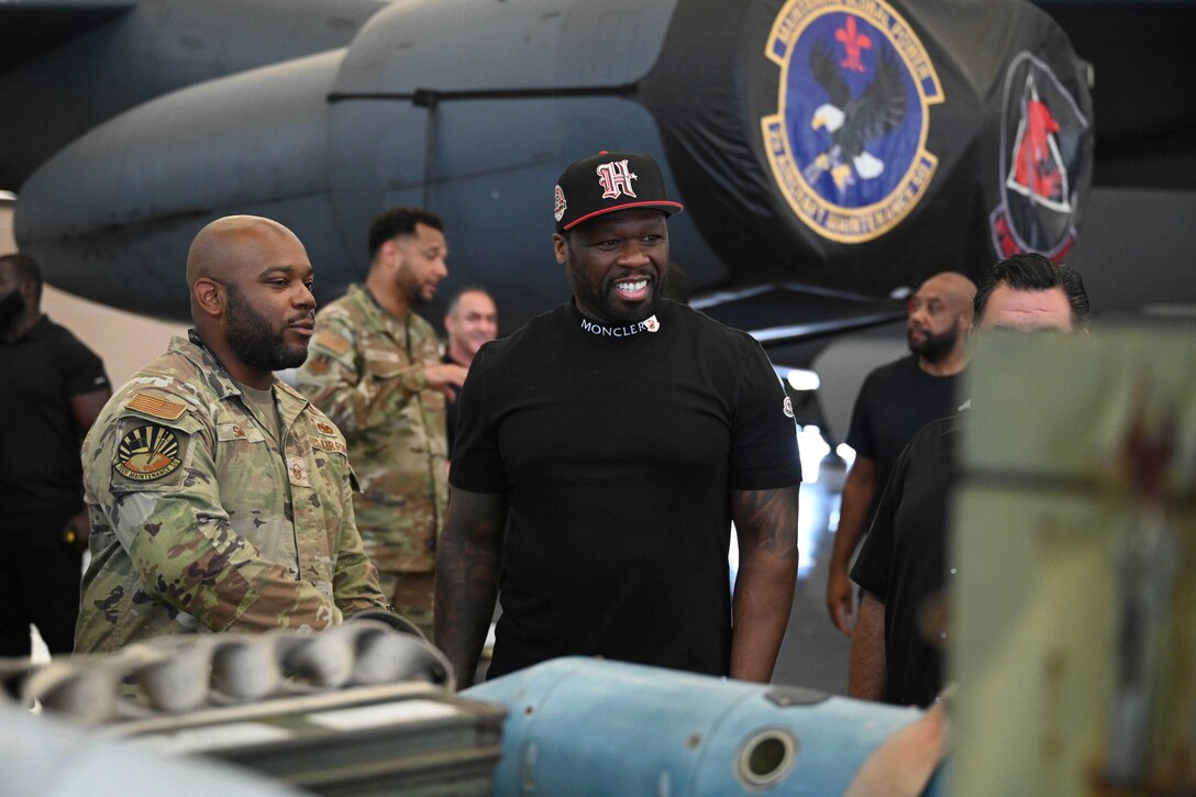 An airman speaks to a person in civilian attire with other service members and a military aircraft in the background.