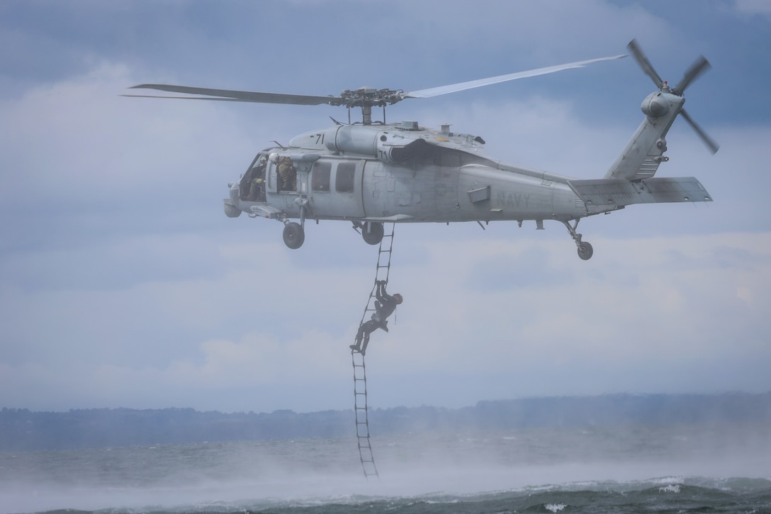 A service member climbs a ladder attached to a helicopter hovering over a body of water.