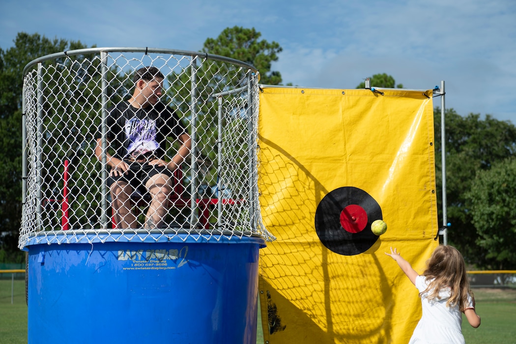 A person sitting on a dunk tank seat and a little girl tossing a softball at the target.