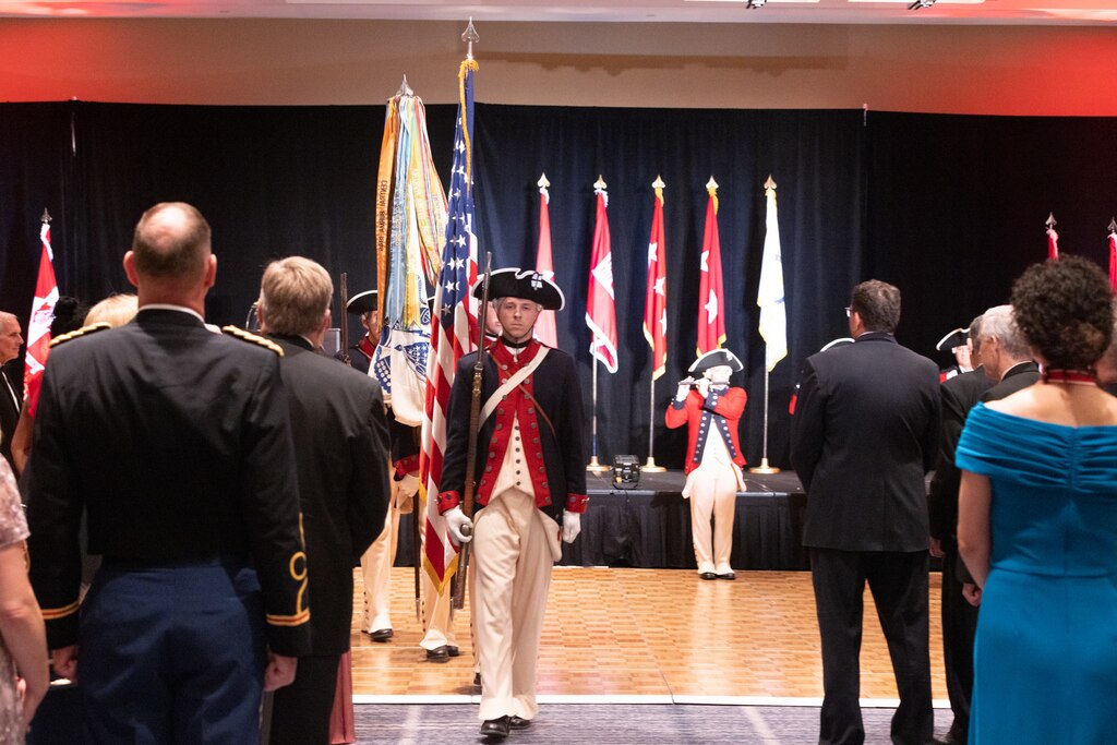 Soldiers in revolutionary era uniforms march out in single file with the American and Army Flags.