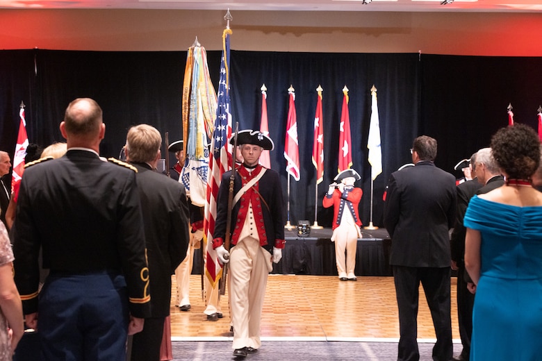 Soldiers in revolutionary era uniforms march out in single file with the American and Army Flags.