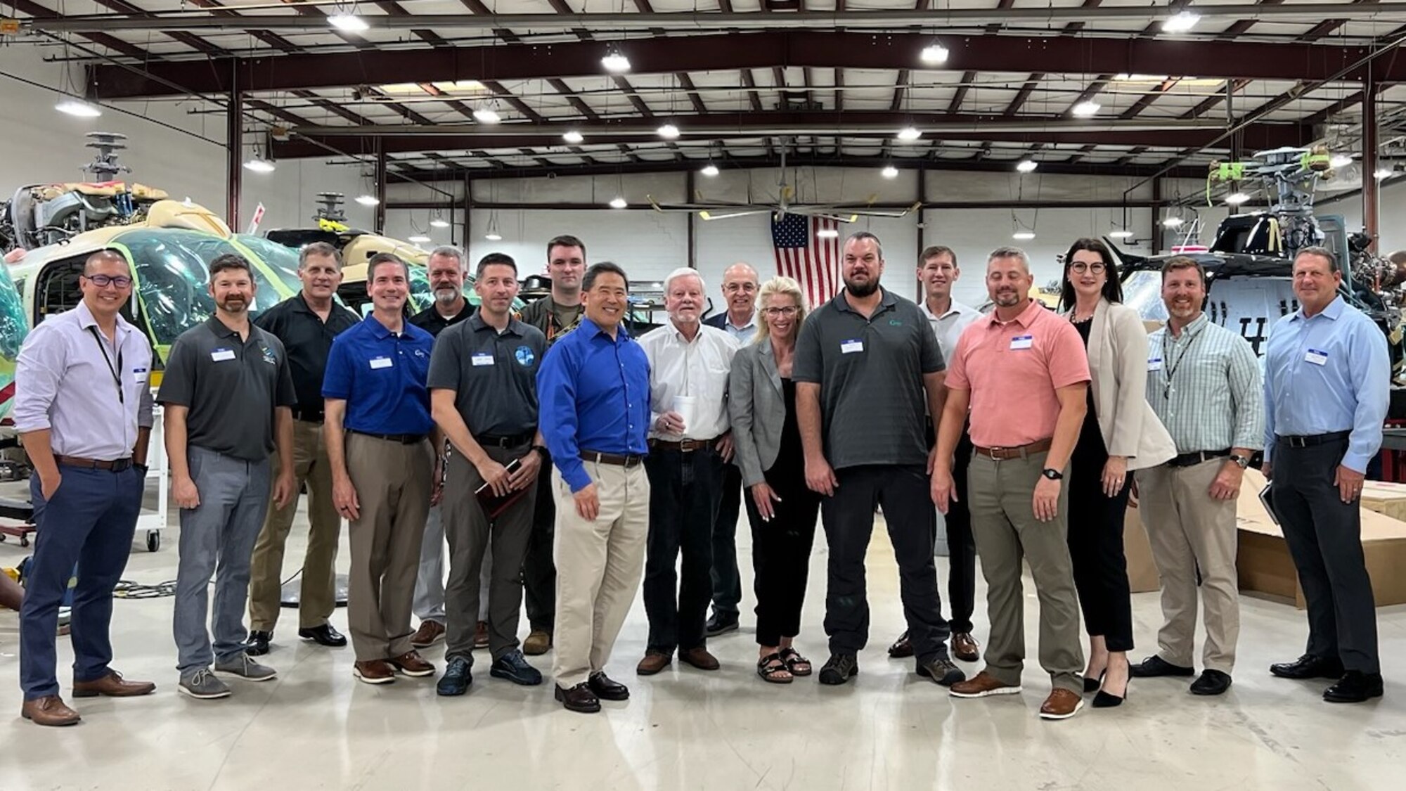Seventeen people stand together to pose and smile for a group photo. They are located inside of an aircraft hangar. Their background shows helicopters in various stages of maintenance with a large American flag on the wall at the very back, which can be seen behind the heads of the people in the group photo.