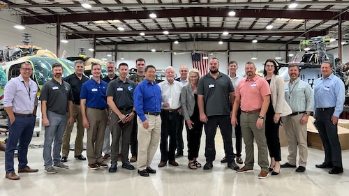 Seventeen people stand together to pose and smile for a group photo. They are located inside of an aircraft hangar. Their background shows helicopters in various stages of maintenance with a large American flag on the wall at the very back, which can be seen behind the heads of the people in the group photo.