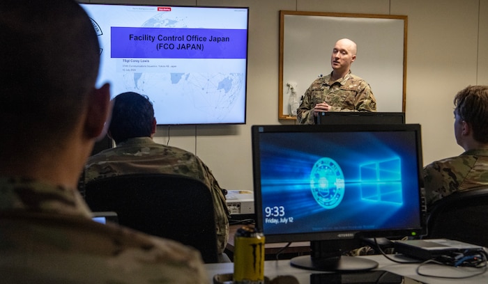A military member presents a slide show in front of a room full of military members sitting behind computer desks.