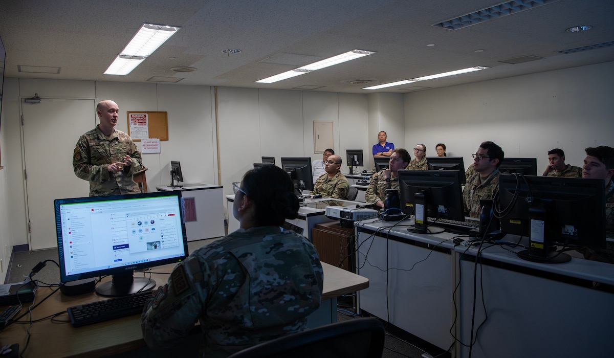 A military member stand in presents in front of a room full of military members sitting behind computer desks.