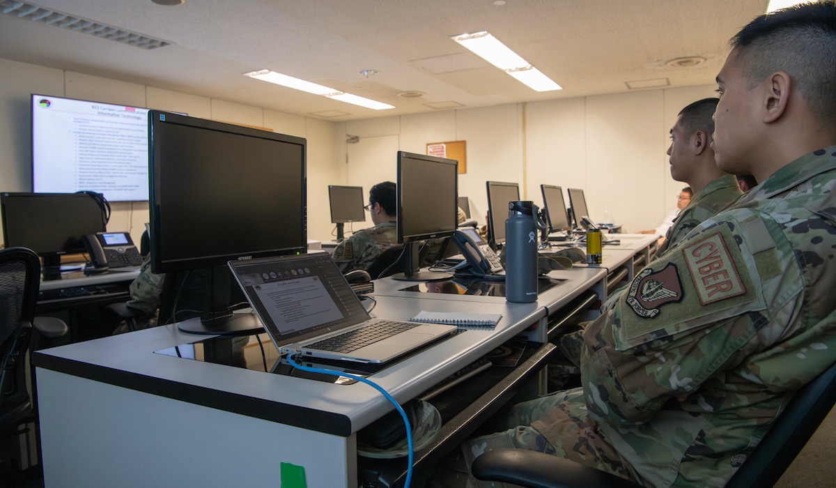 Military members look at a screen at the front of the room while sitting behind computer desks.