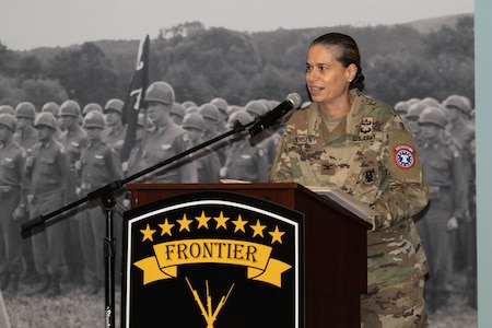 Female Soldier in uniform behind a podium with a brigade logo, in front of a General George Patton leadership mural