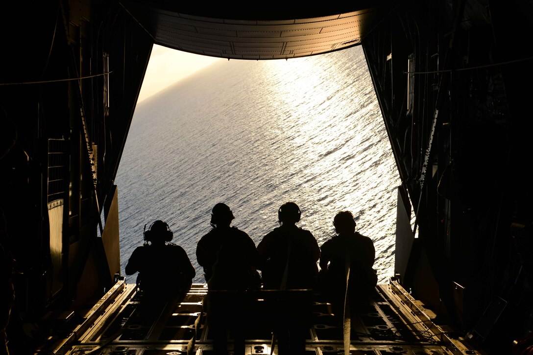 Four airmen are silhouetted against a sunlit sea as they look down from the back of an aircraft.