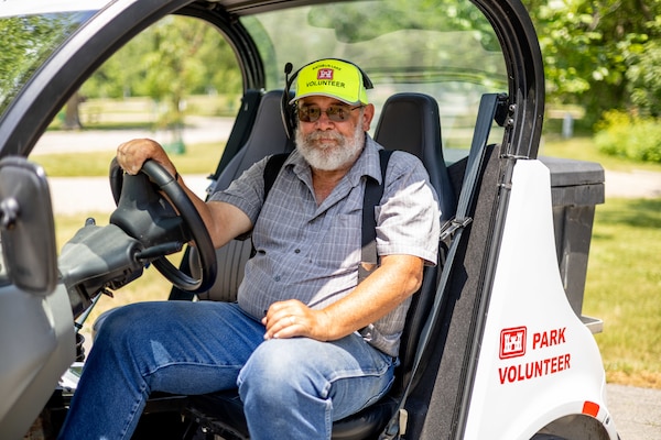 A man in blue jeans, a blue shirt, and a yellow hat sits in a golf cart with grass and trees in the background.