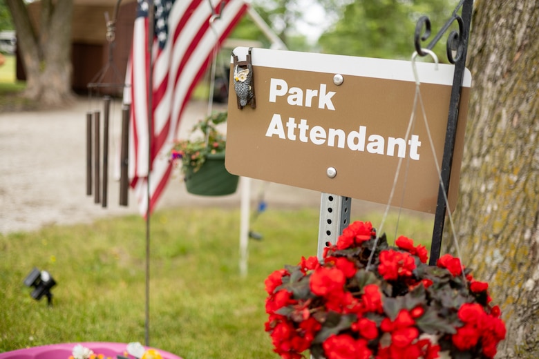 A brown sign reads park attendant with an American flag in the background and red flowers in the foreground.