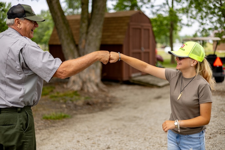 A man in a grey shirt and green pants fist bumps a girl in a brown shirt and a yellow hat.