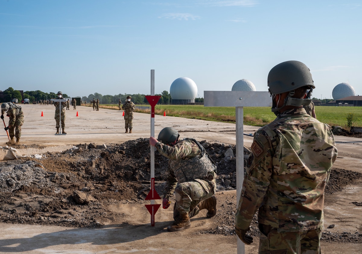 U.S. Airmen in uniform and helmets work on a construction site, setting up measuring equipment near a man made crater.