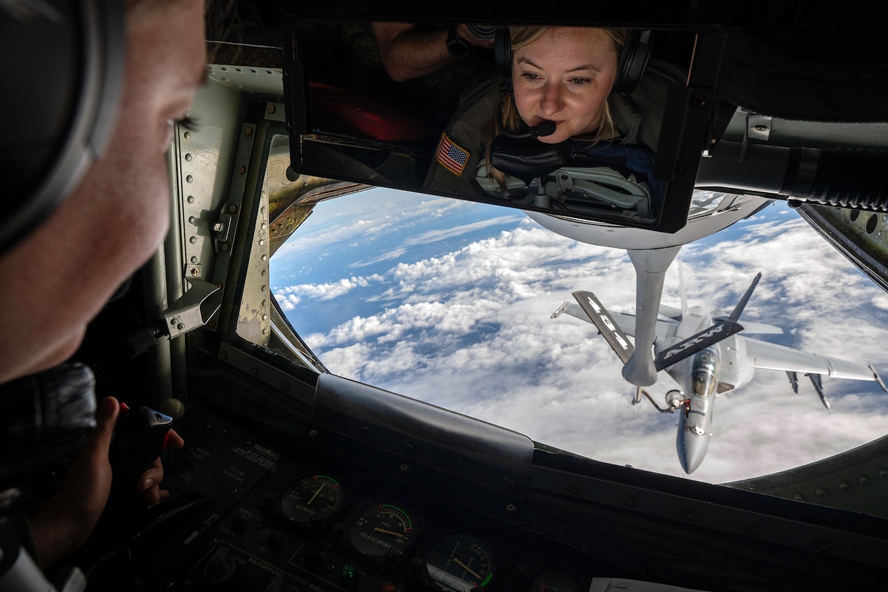 A technician facing a mirror and its reflection looks out of a window of an aircraft at another aircraft being refueled high above the clouds.