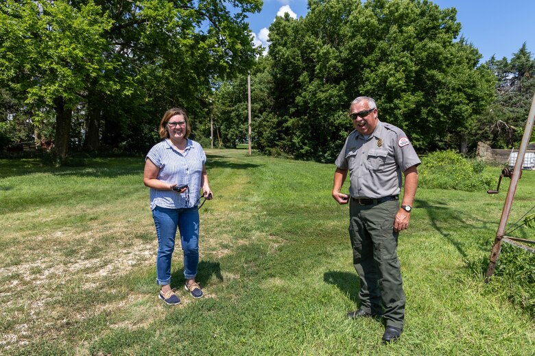 A woman in blue jeans and a blue striped top and a man in a grey shirt and green pants laugh while standing in a grassy area with trees in the background.