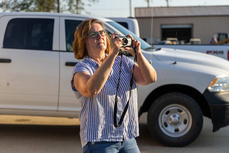 A woman in blue jeans and a blue striped top holds a camera in front of a white truck.