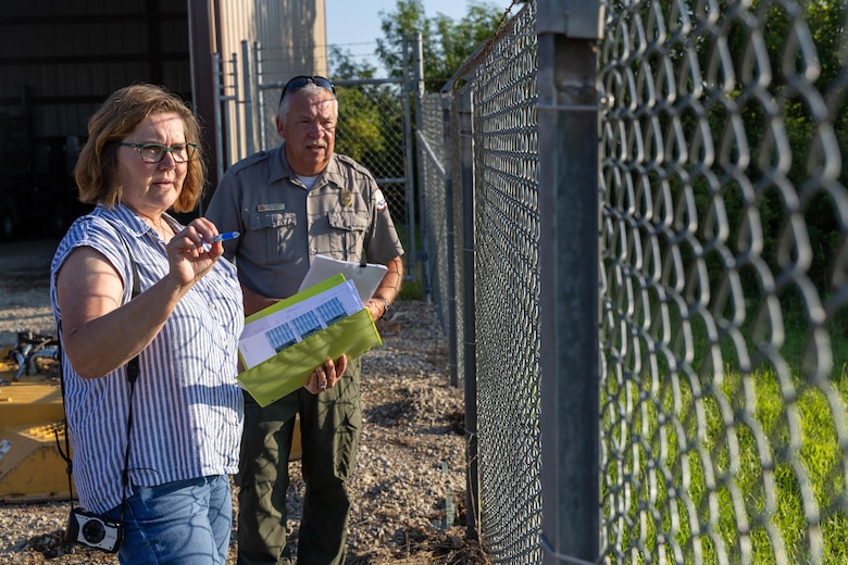 A man in a grey shirt and green pants and a woman in blue jeans and a blue striped shirt holding a green binder points at a chain link fence with grass on the right hand side.