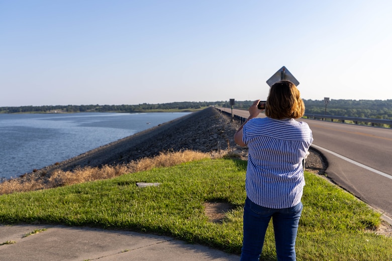 A woman in a blue striped shirt holds a camera while taking a picture of a road with blue water and grass on the left and blue sky in the background.