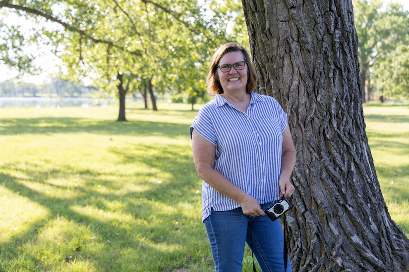 A woman in blue jeans and a blue striped shirt holds a camera while smiling in front of a tree with grass in the background.