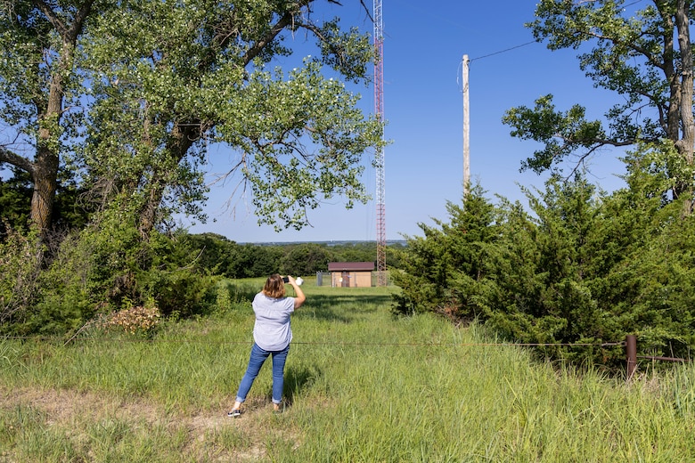A woman in blue jeans and a blue striped shirt takes a photo of a brown building with trees, grass, and blue sky in the background.
