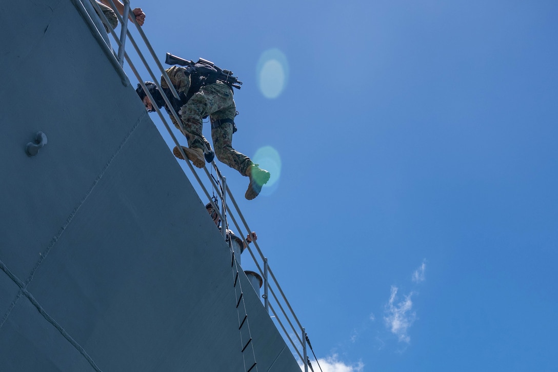 A sailor climbs the side of a ship during training as others watch.