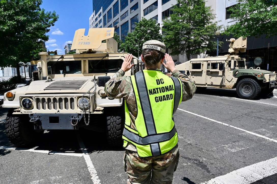 A service member wearing a reflective vest that reads "DC National Guard" raises their hands to direct military vehicles on a city street.