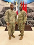 Two Army Soldiers in uniform stand in front of a flag display.