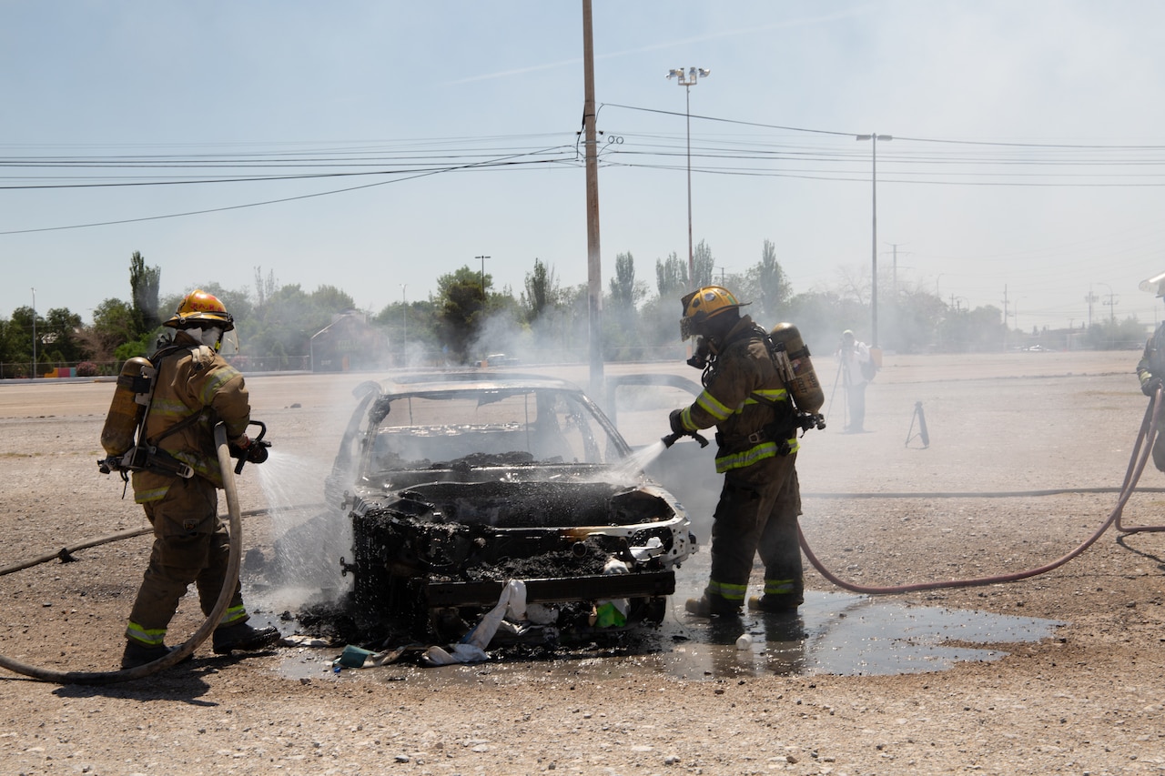 Two people wearing firefighter gear extinguish a vehicle fire in a flat field.
