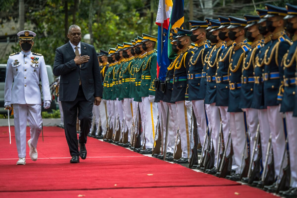 A person wearing a business suit holds their hand over their heart while walking near a formation of foreign service members.