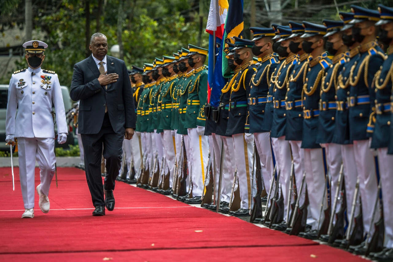A person wearing a business suit holds their hand over their heart while walking near a formation of foreign service members.