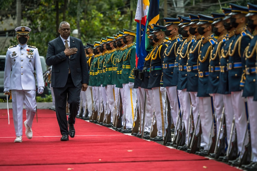 A person wearing a business suit holds their hand over their heart while walking near a formation of foreign service members.