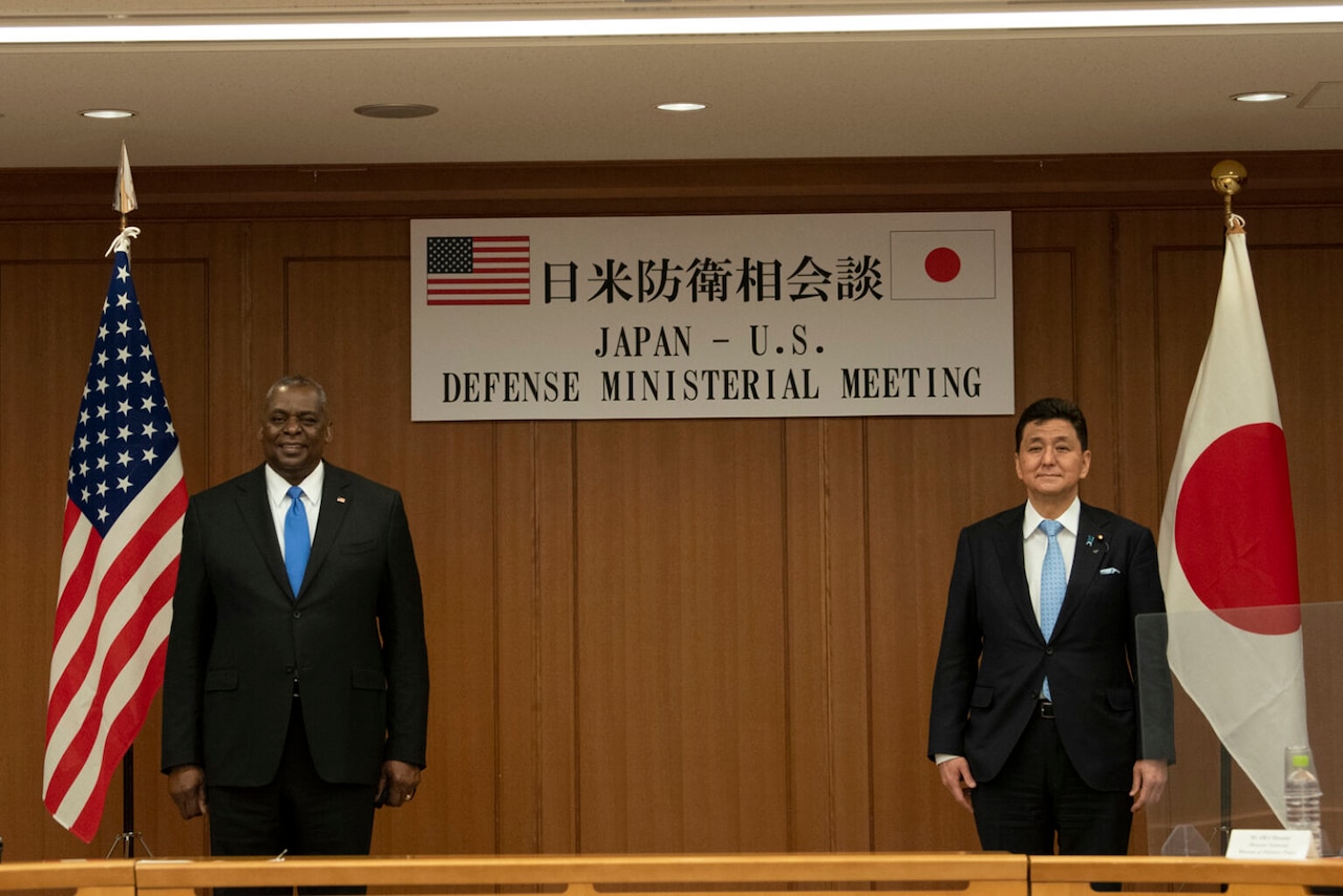 Two people wearing business suits stand in front of U.S. and Japanese flags.